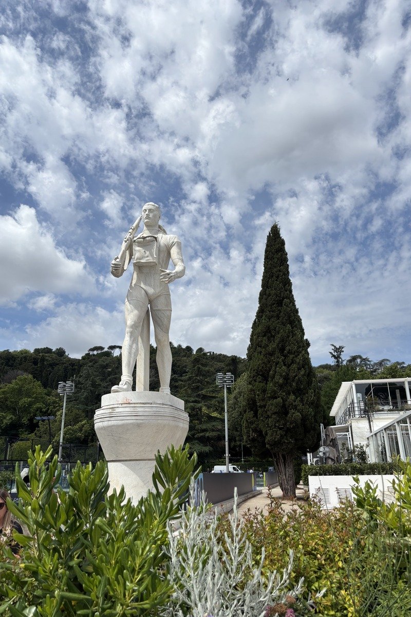 Statue blanche d'un joueur de tennis sur un piédestal entouré de verdure