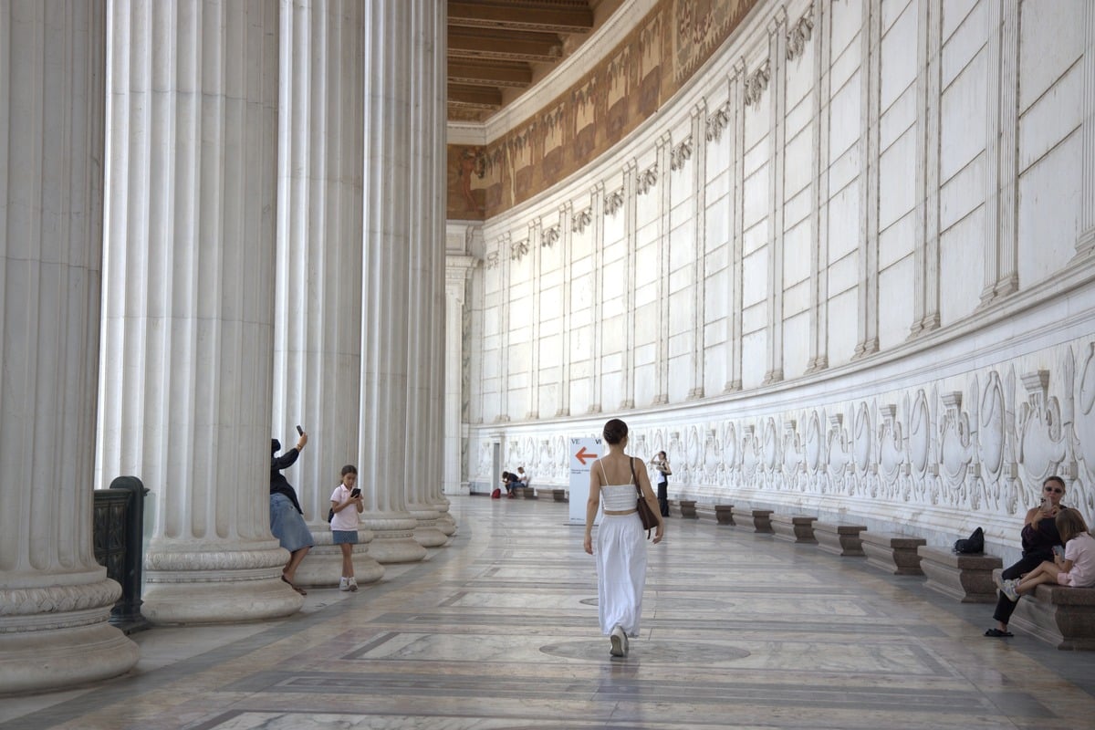 Femme en blanc marchant sous une colonnade de marbre avec visiteurs