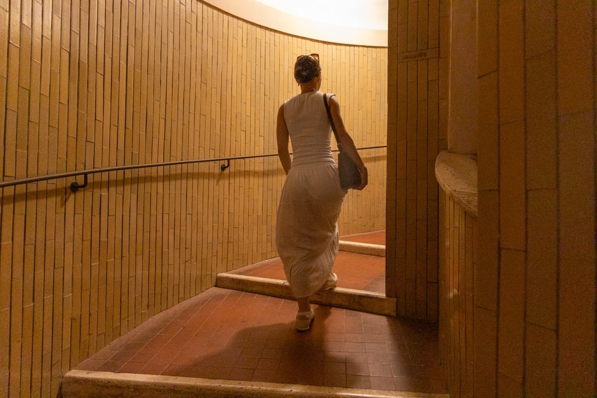 Femme en robe blanche montant un escalier aux murs carrelés ocre