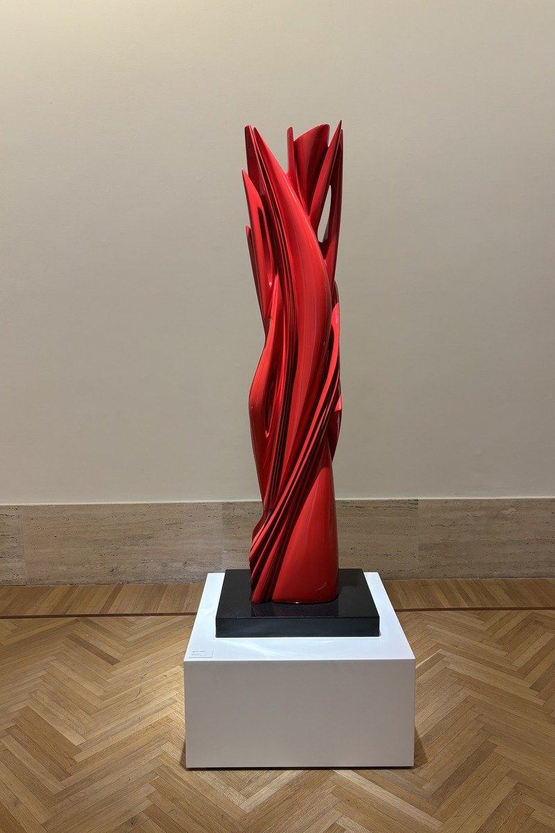 Sculpture abstraite rouge vif sur socle blanc, formes torsadées élancées